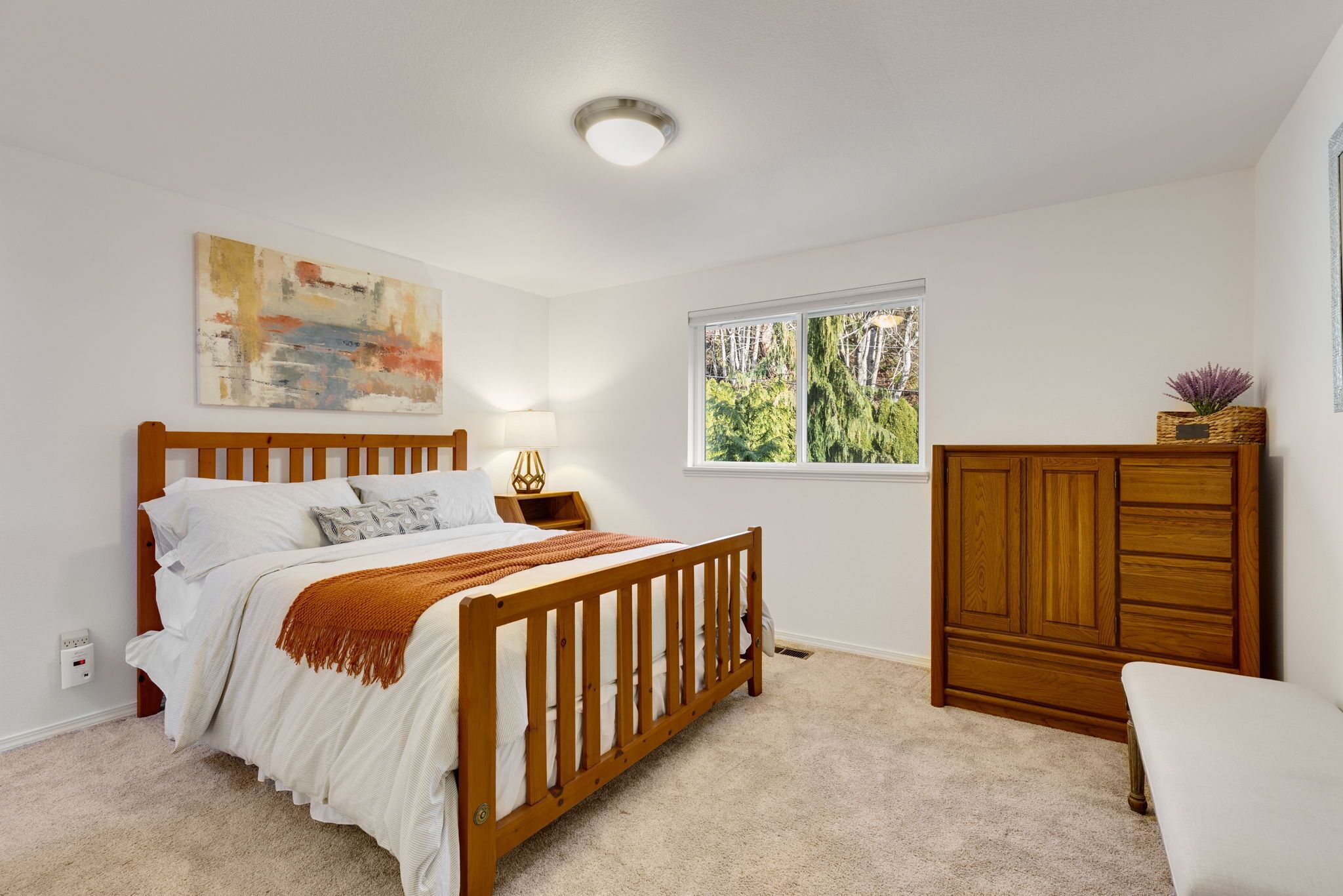 Un dormitorio con una cama de madera, una mesita de noche, un aparador, una ventana con vista a árboles y una lámpara de techo.