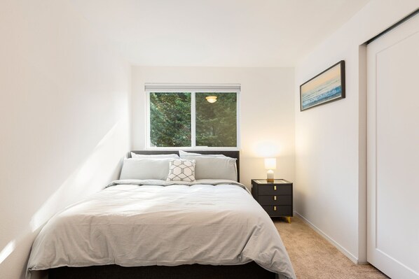 A bedroom with a bed, a nightstand, a lamp, a window, and a painting on the wall.