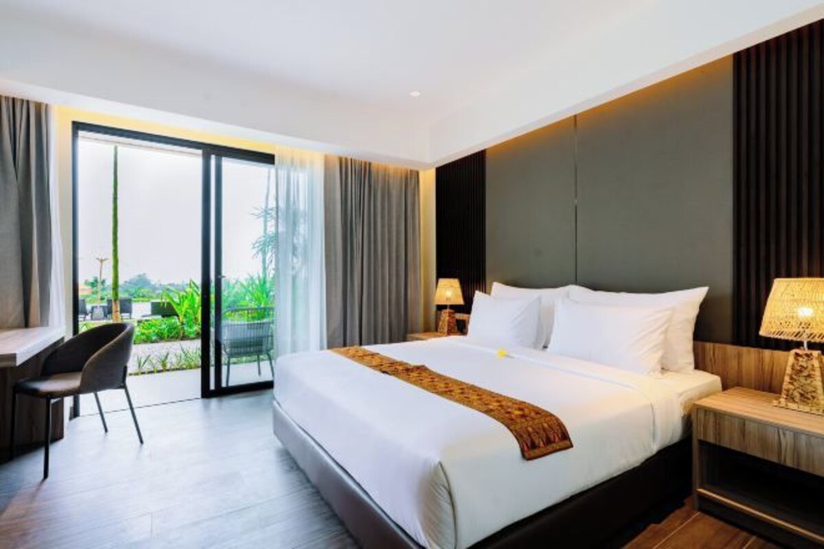 A modern hotel room with a large bed, a desk, and a view of greenery through the sliding glass door.