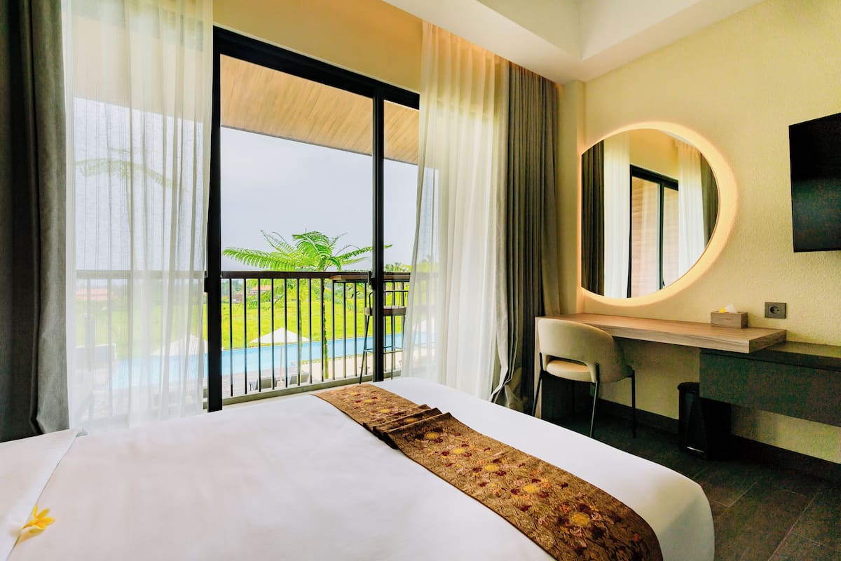 A hotel room with a bed, a desk, a chair, a round mirror, and a view of a pool and greenery.