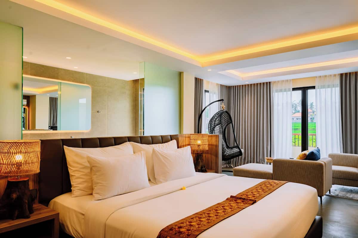 A hotel room with a large bed, a sitting area with a chair, a small table, and a large window with curtains.