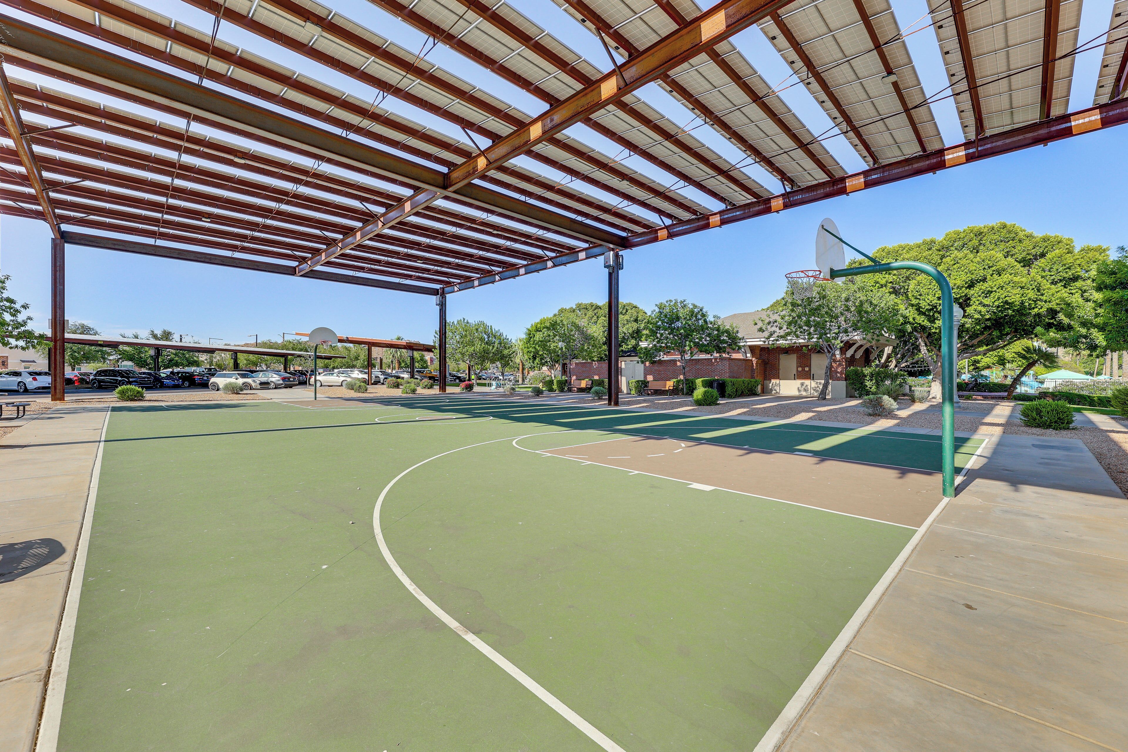 An outdoor basketball court with a metal roof structure, surrounded by trees and a parking area.