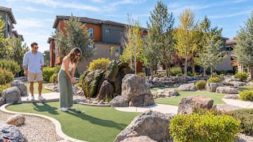 Een koppel speelt golf in een verzorgd buitengebied met een putting green, omringd door rotsen en bomen, met woongebouwen op de achtergrond.