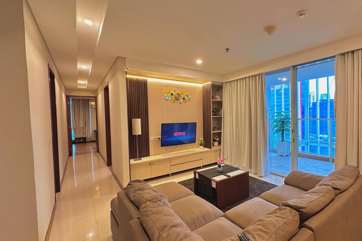 A modern living room with a flat-screen TV, a sofa, a coffee table, and a large window with curtains.