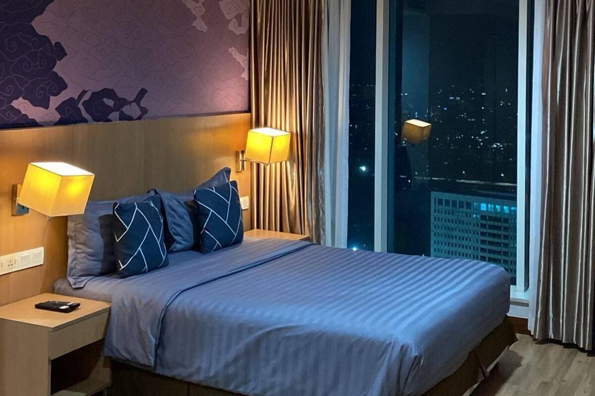 A modern hotel room with a large bed, two bedside lamps, a nightstand, and a view of the city at night.