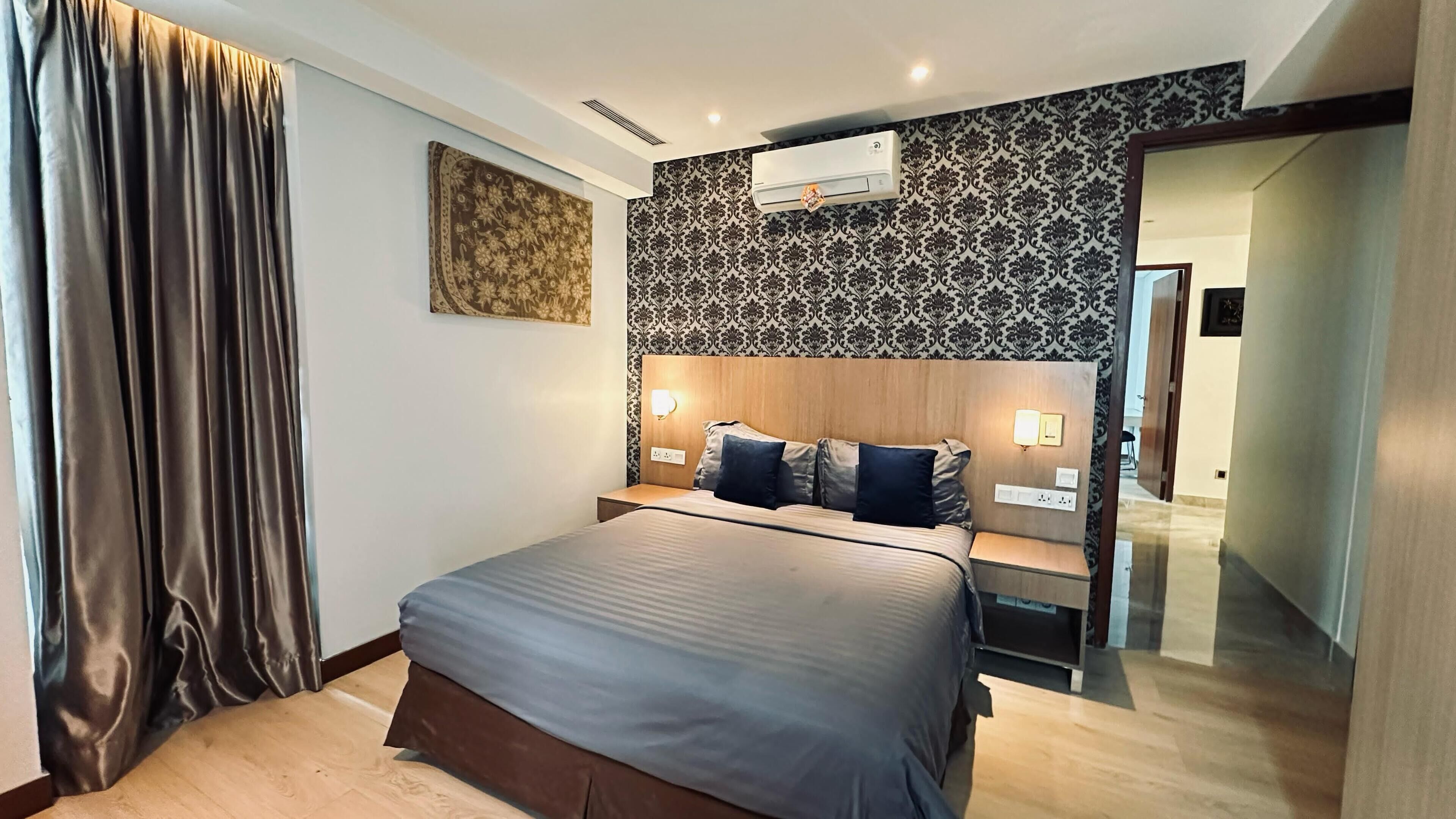 A modern hotel room with a large bed, a headboard with integrated lighting, a decorative wall panel, and an air conditioning unit.