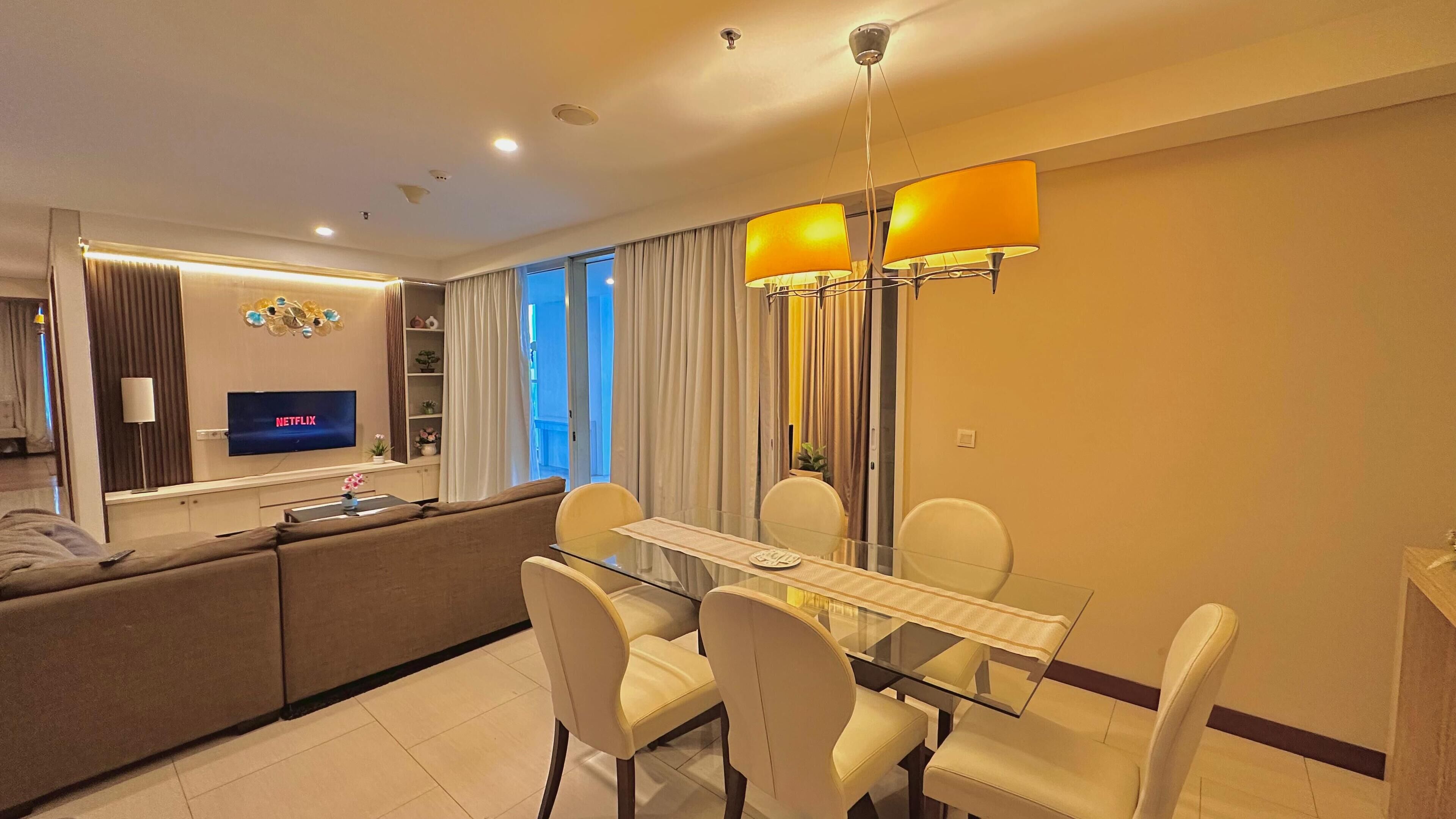 Una sala de estar moderna con una mesa de comedor de vidrio, sillas blancas, un sofá, un televisor que muestra Netflix y un ventanal con cortinas.