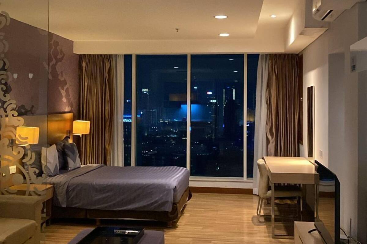 A modern hotel room with a large bed, a sofa, a desk, and a TV. The room has a city view through large windows.