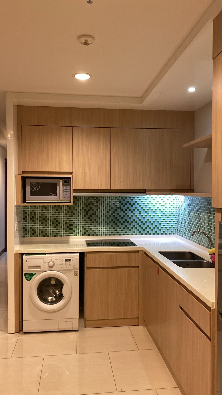A compact kitchen with wooden cabinets, a microwave, a washing machine, and a tiled backsplash.