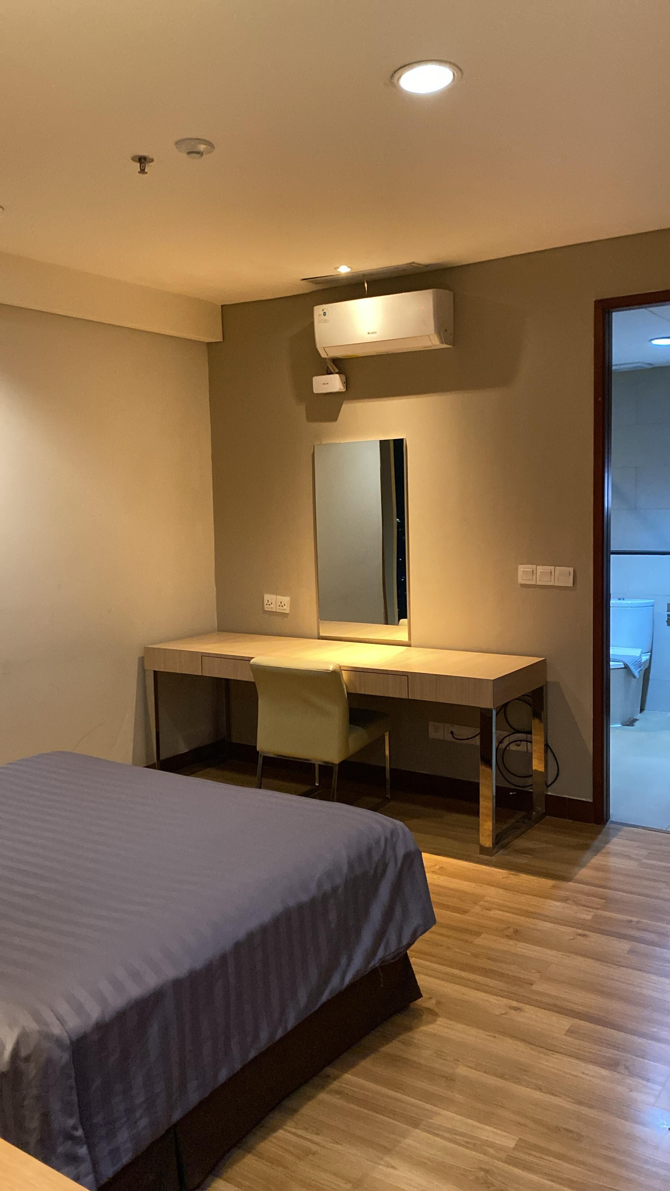 A bedroom with a bed, a desk, a chair, a mirror, and an air conditioning unit.