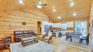 Un amplio salón con techo de madera, un sofá de cuero marrón, una mesa de comedor con sillas y una cocina con armarios blancos y un refrigerador.