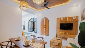 A dining area with a wooden table set for two, a television mounted on a wooden panel wall, and a large plant in the corner.