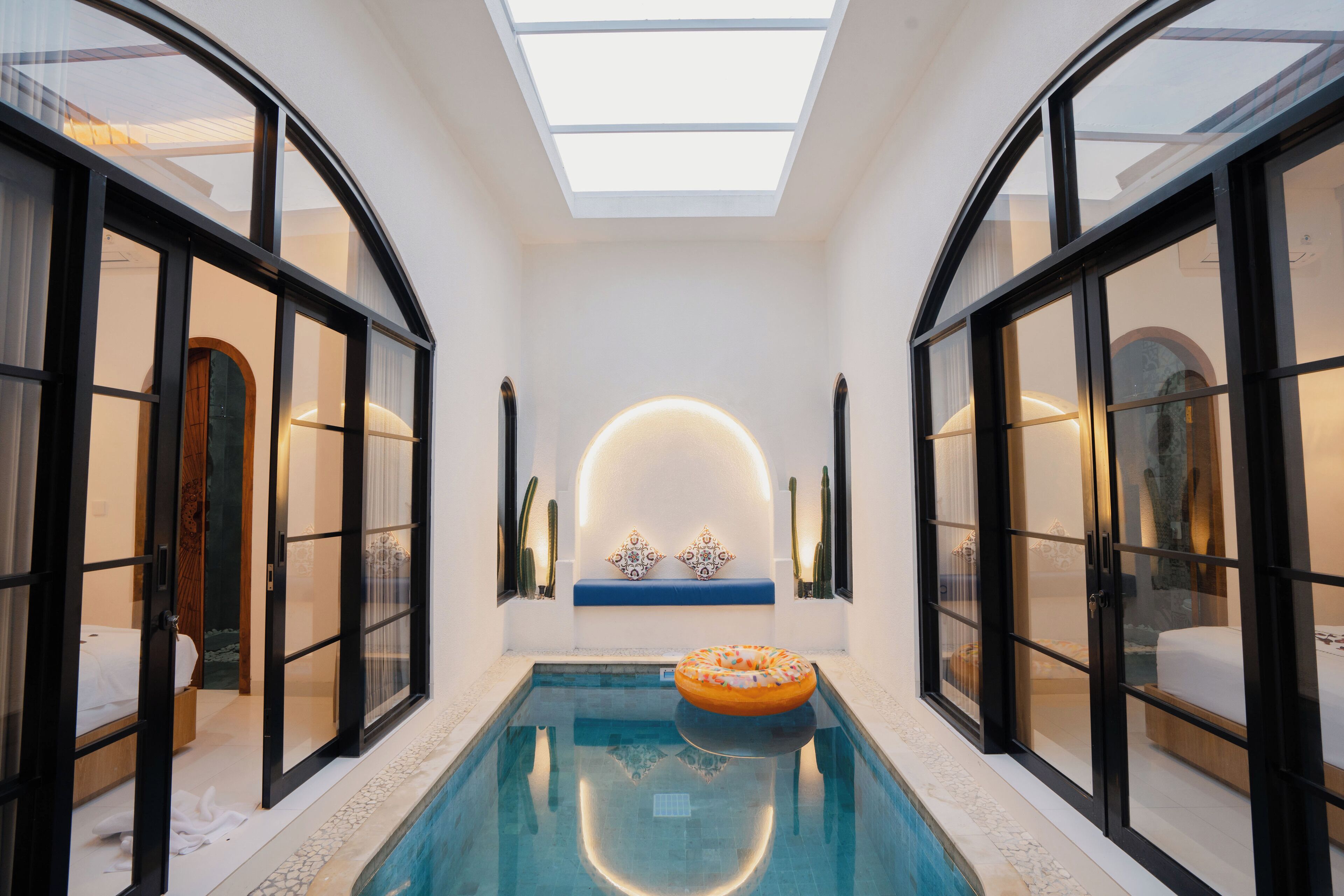 A modern indoor pool with glass walls and a skylight, a floating ring, and a decorative wall feature.