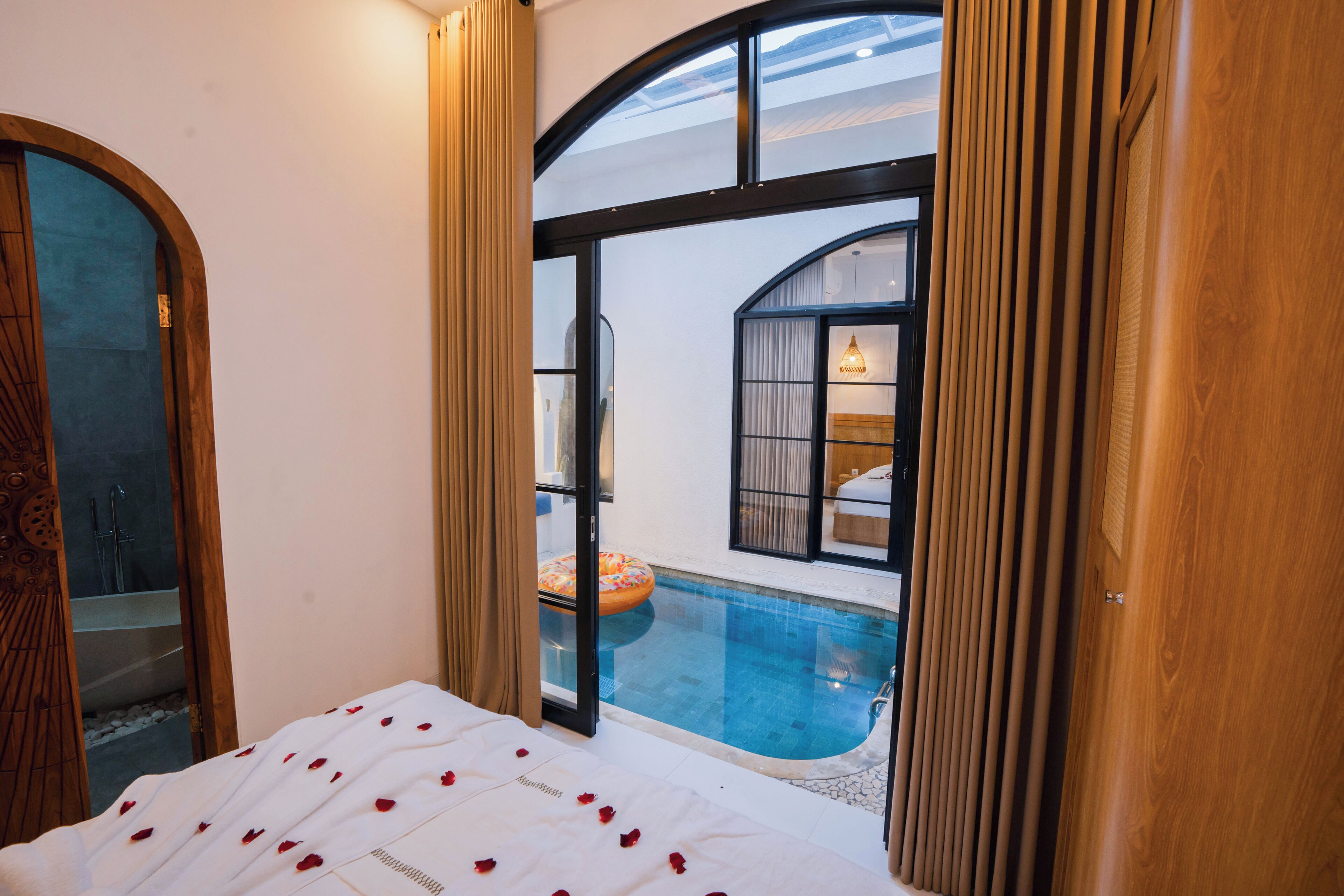 Een slaapkamer met een groot raam dat uitkijkt op een zwembad, een bed met rozenblaadjes en zicht op een badkamer met een bad.