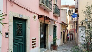 Una estrecha calle empedrada con edificios de arquitectura tradicional, incluyendo un edificio rosa con puerta verde y un letrero que indica un comercio.