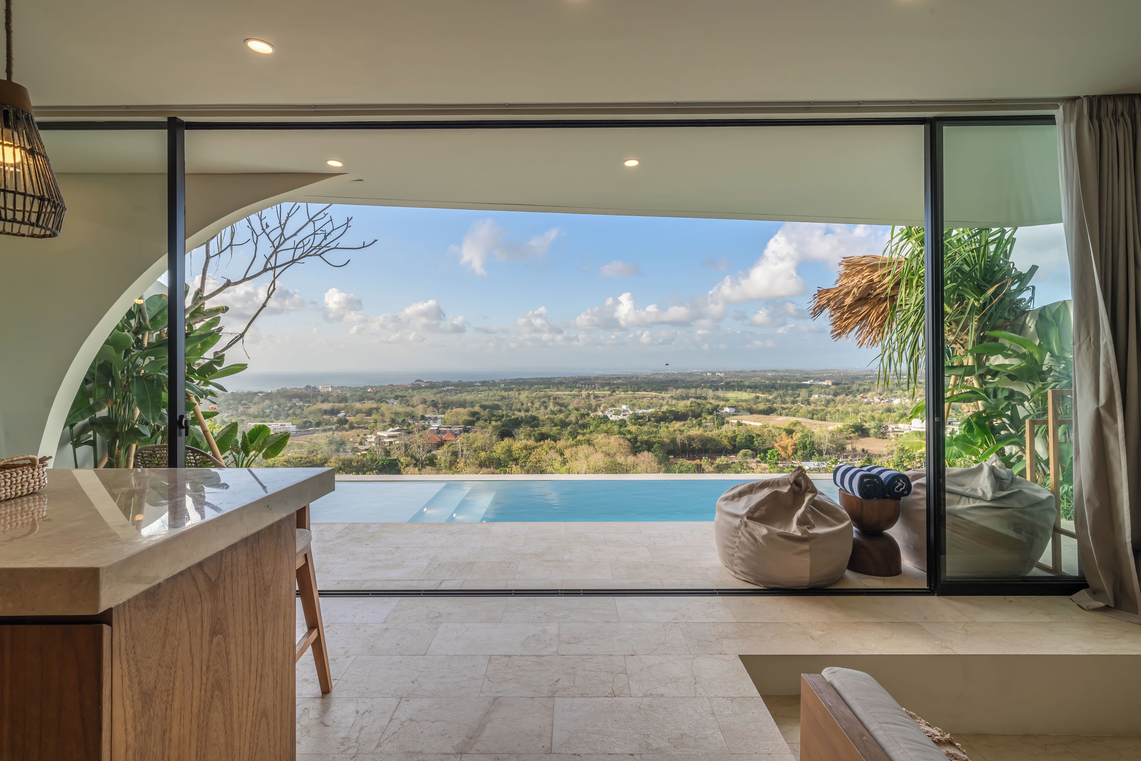 A modern room with a large sliding glass door leading to a pool, a wooden bar area, and a view of a lush landscape.