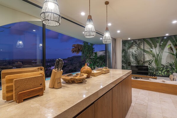 Une cuisine moderne dotée d’un grand îlot central, d’ustensiles en bois et offrant une vue sur la ville au crépuscule.