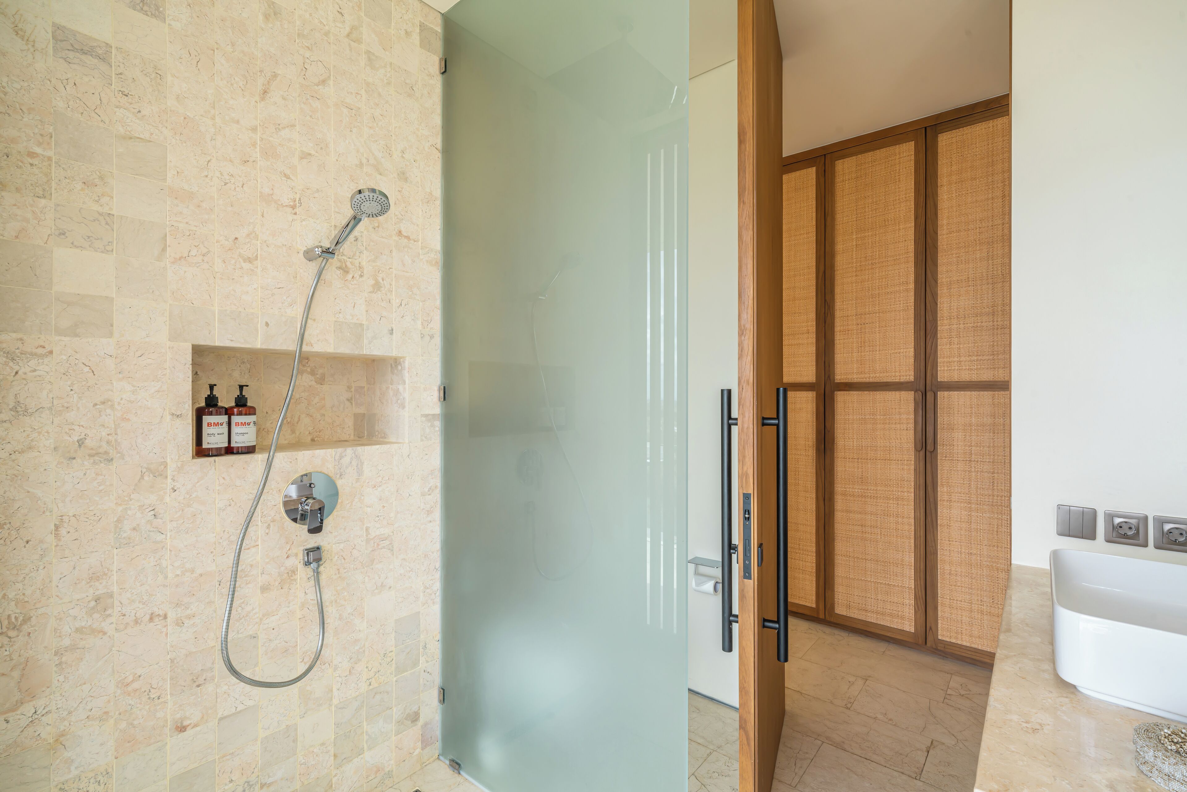 A modern bathroom with a glass shower enclosure, a built-in shelf with toiletries, and a wooden door leading to another room.