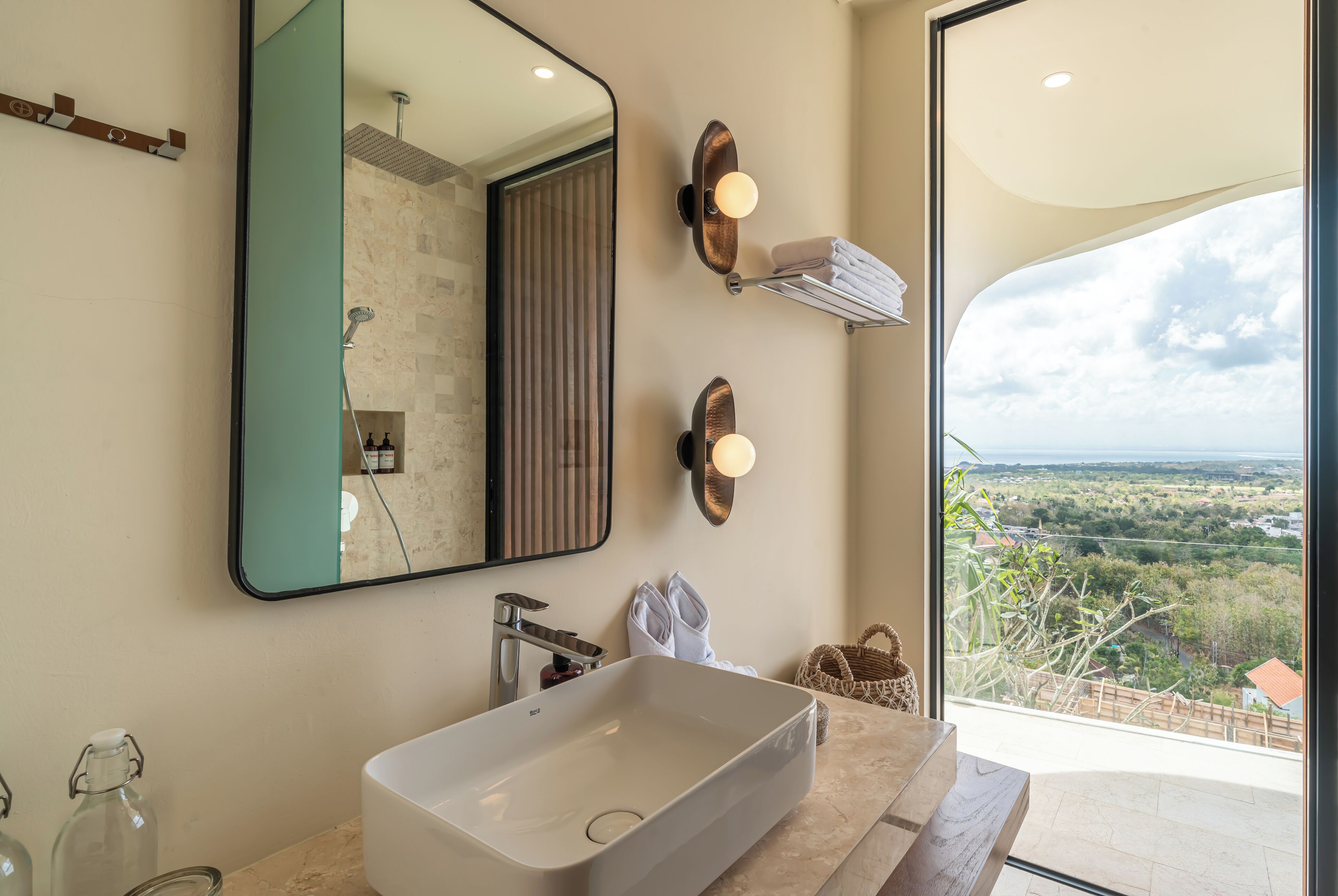 A bathroom with a large mirror, a white sink, and a view of the countryside through a large window.