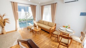 A modern living room with a sofa, dining table, and chairs. There is a large window with curtains, a potted plant, and an air conditioning unit on the wall. - Mandala Hill Private Pool Villa Estate Nusa Dua (Nusa Dua)