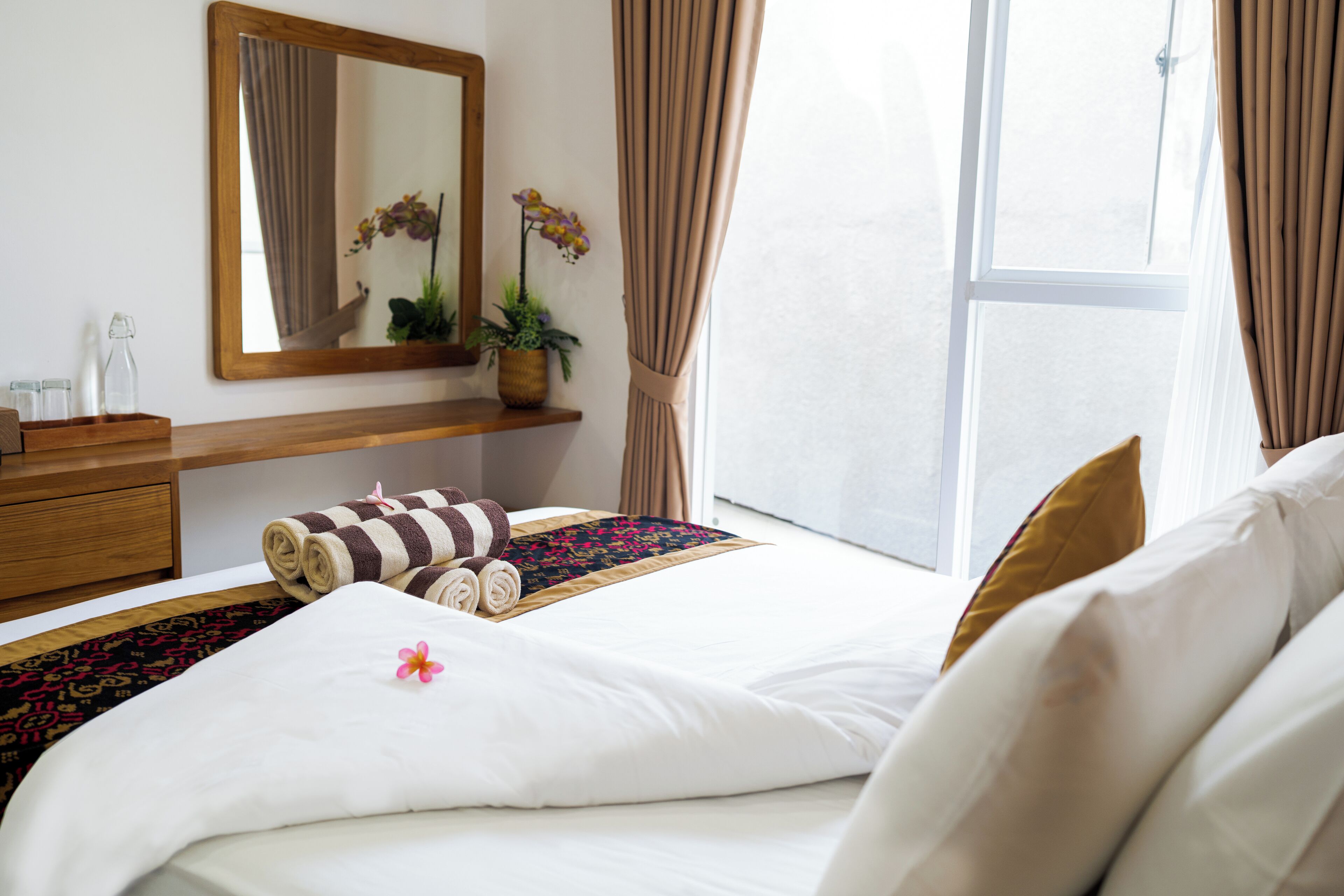 A hotel room with a bed, a wooden dresser, a mirror, a window with curtains, and a potted plant.