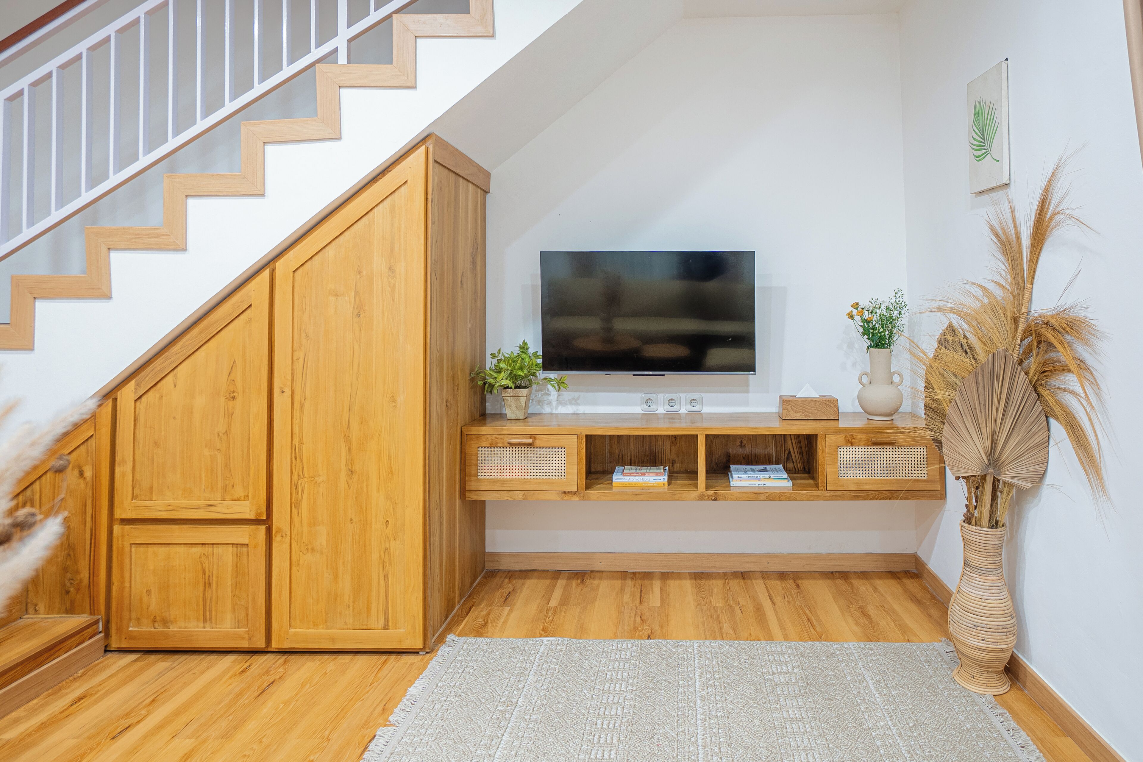 Una escalera con escalones de madera y barandilla blanca, que conduce a una plataforma con una televisión de pantalla plana, armarios de madera y una planta decorativa.