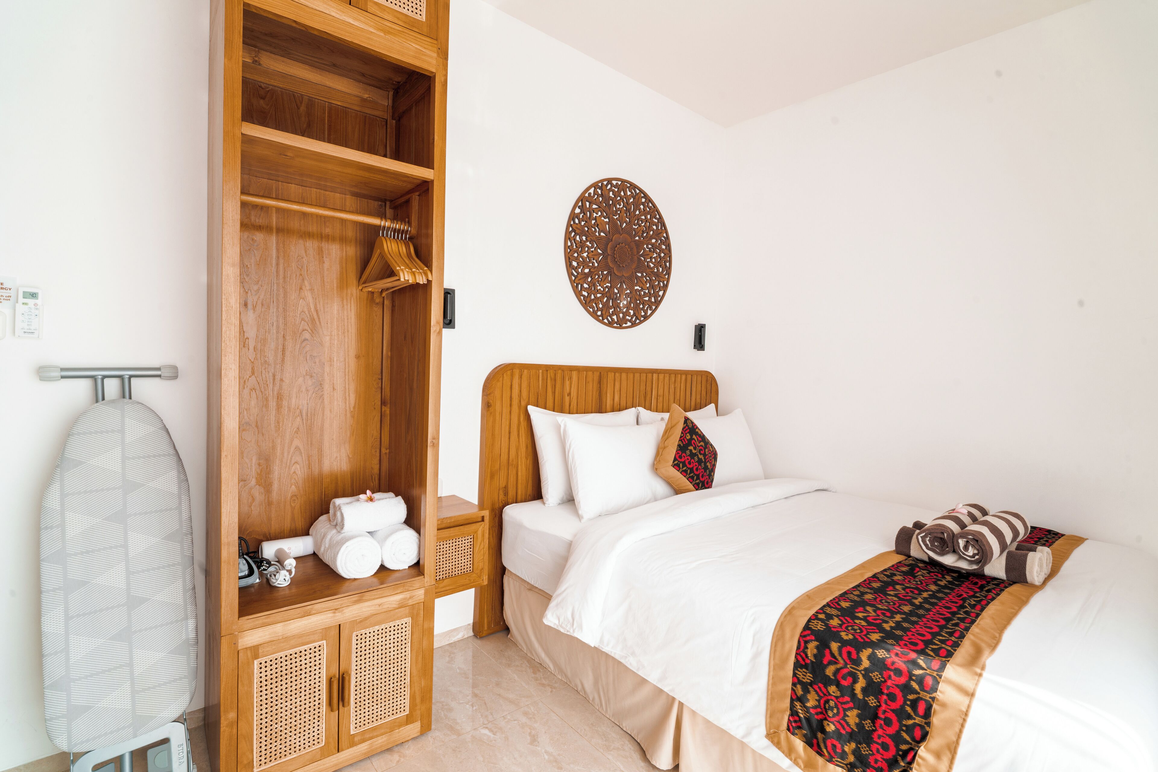 A hotel room with a wooden wardrobe, a bed with white bedding, a decorative wall piece, and a towel rack.