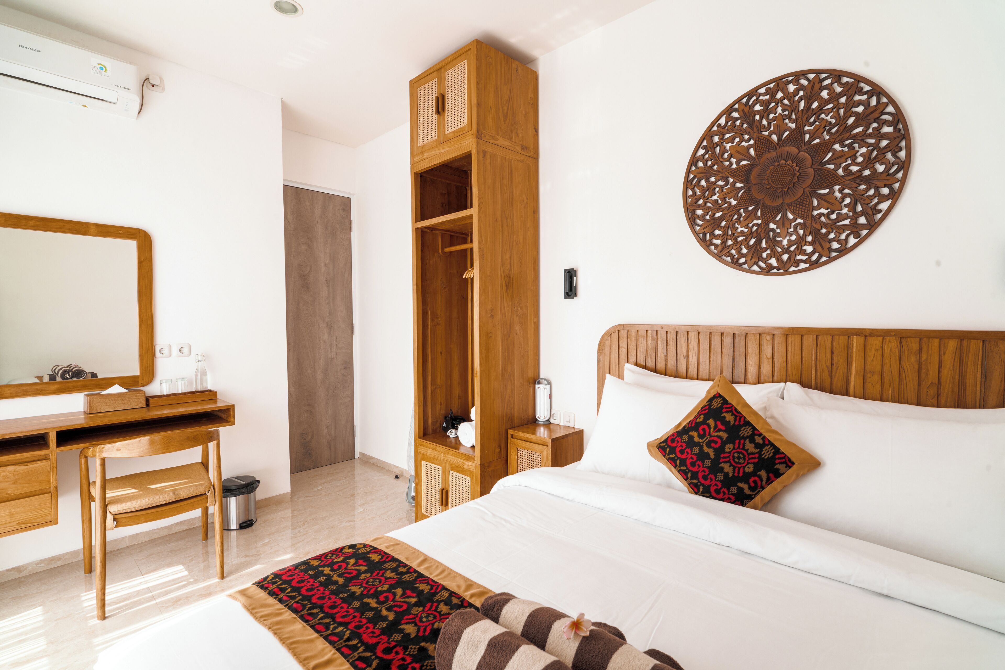A hotel room with a wooden desk, a wardrobe, a bed with white bedding, and a decorative wall piece.