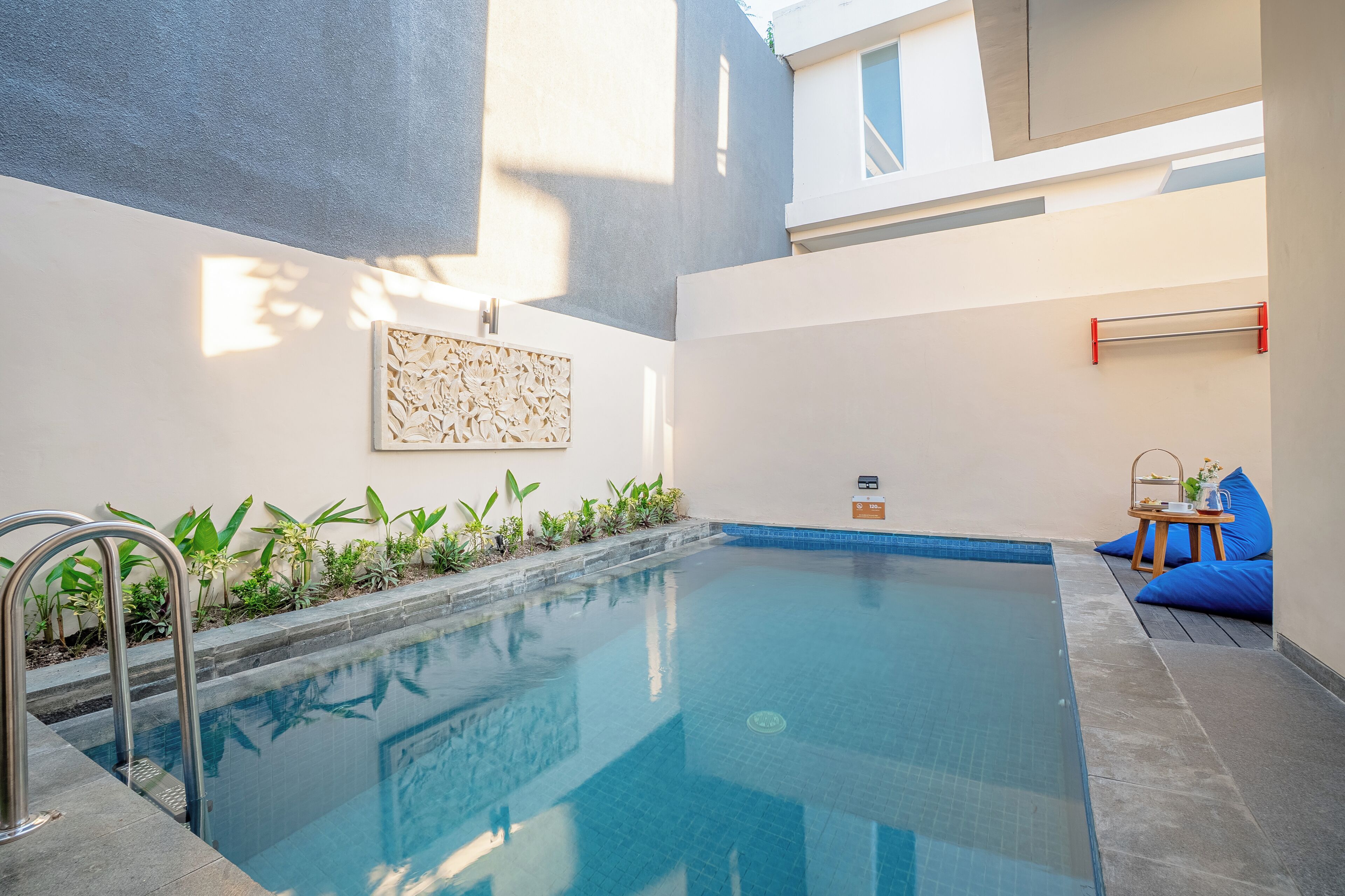 Una piscina con una escalera metálica, rodeada de plantas y un pequeño espacio de estar con una mesa y cojines.