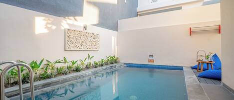 Una piscina con una escalera metálica, rodeada de plantas y un pequeño espacio de estar con una mesa y cojines.