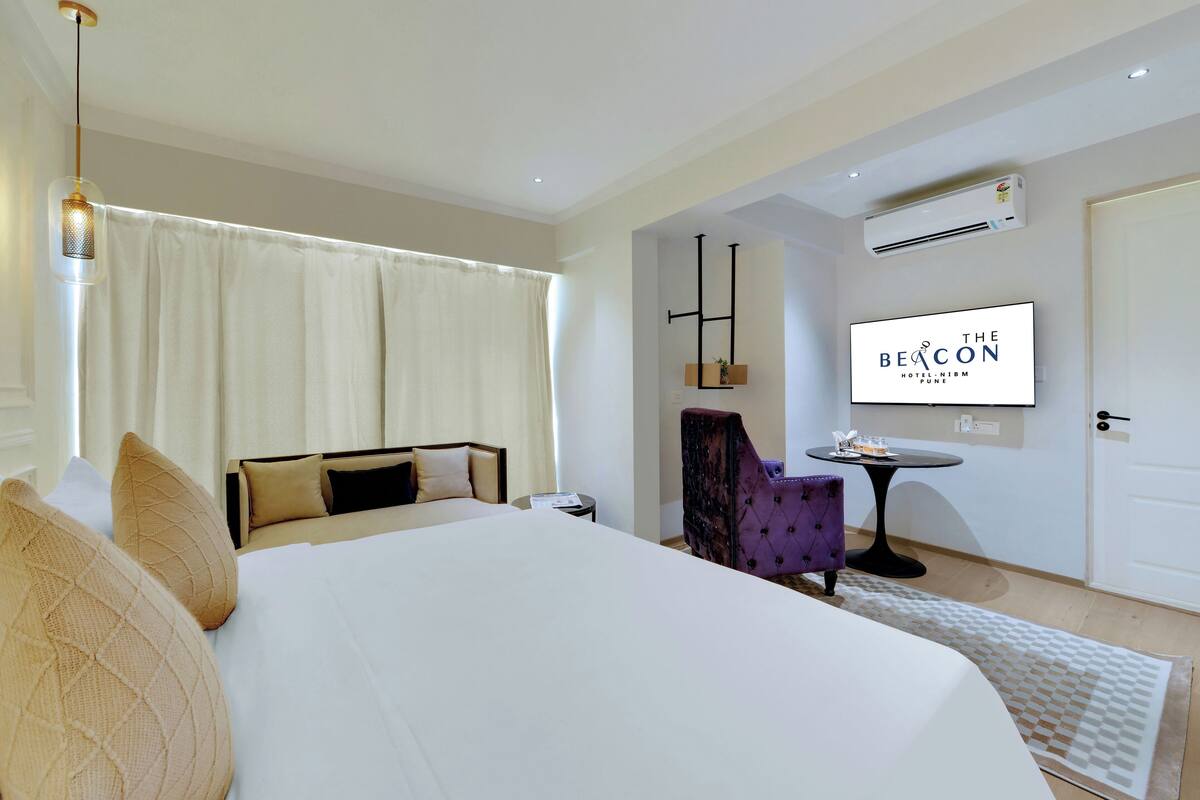 Une chambre d’hôtel avec un grand lit, une petite table, un fauteuil violet et un téléviseur fixé au mur qui diffuse une image de « The Beacon Hotel, Mumbai ».