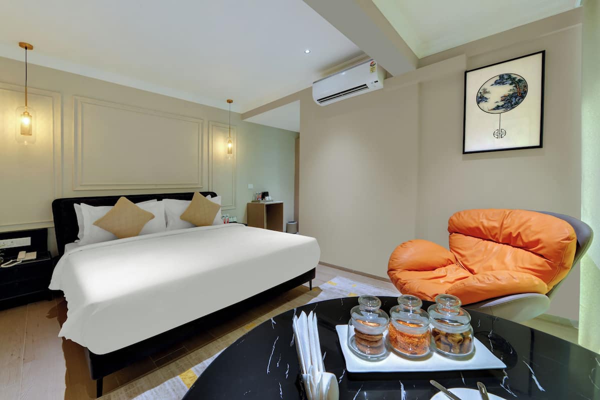 Une chambre d’hôtel avec un lit, un fauteuil orange, une table à manger sur laquelle se trouve un plateau de collations, et une œuvre d’art encadrée accrochée au mur.