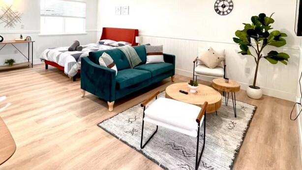 Una sala de estar moderna y bien iluminada con un sofá azul verdoso, una mesa de centro redonda y una mesita auxiliar. Se observa una cama con cabecera blanca y una ventana con persianas. En la pared hay un reloj, y una planta en maceta es visible.