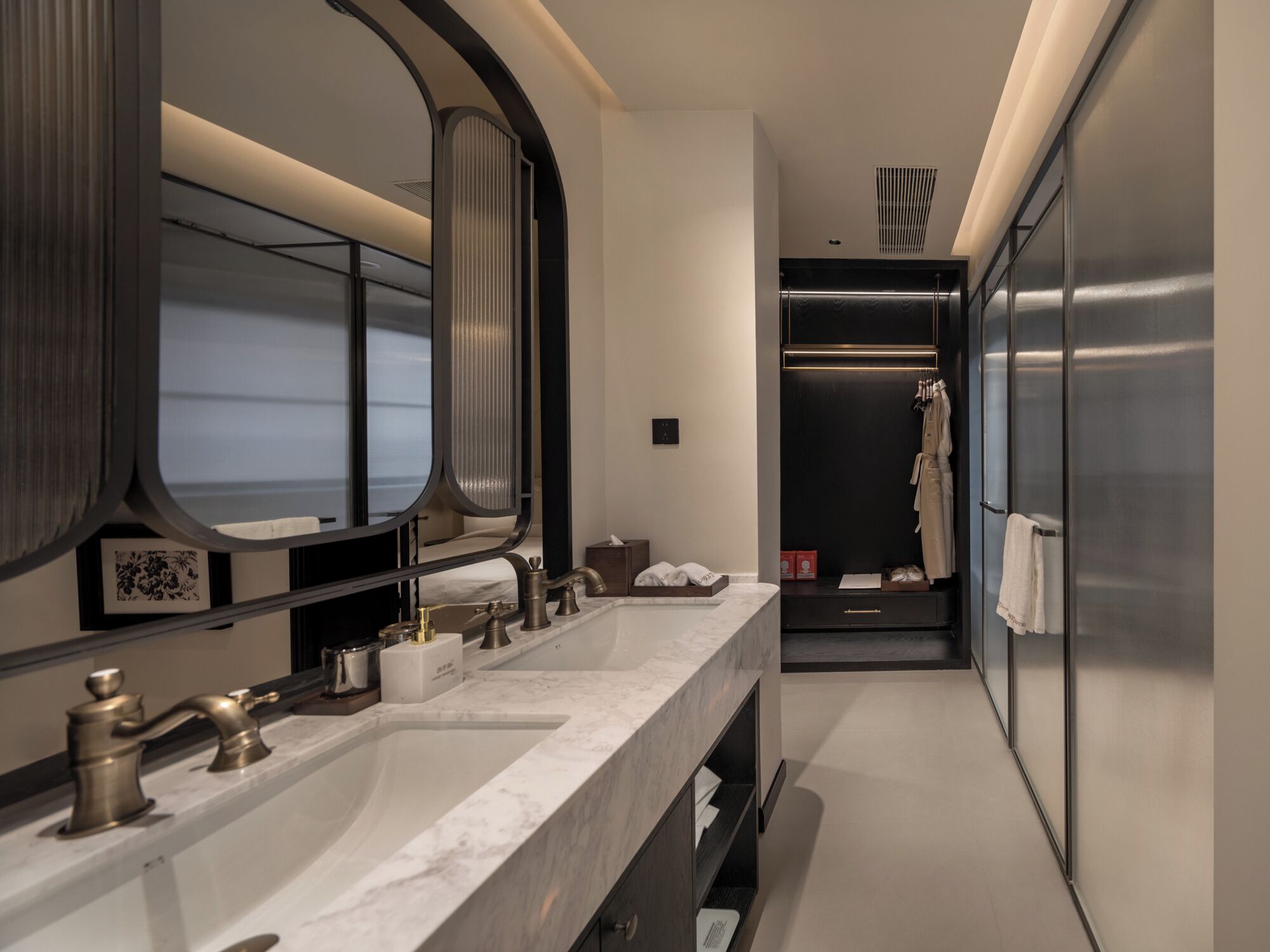 A modern bathroom with a large mirror, two sinks, a walk-in closet, and a stainless steel refrigerator.