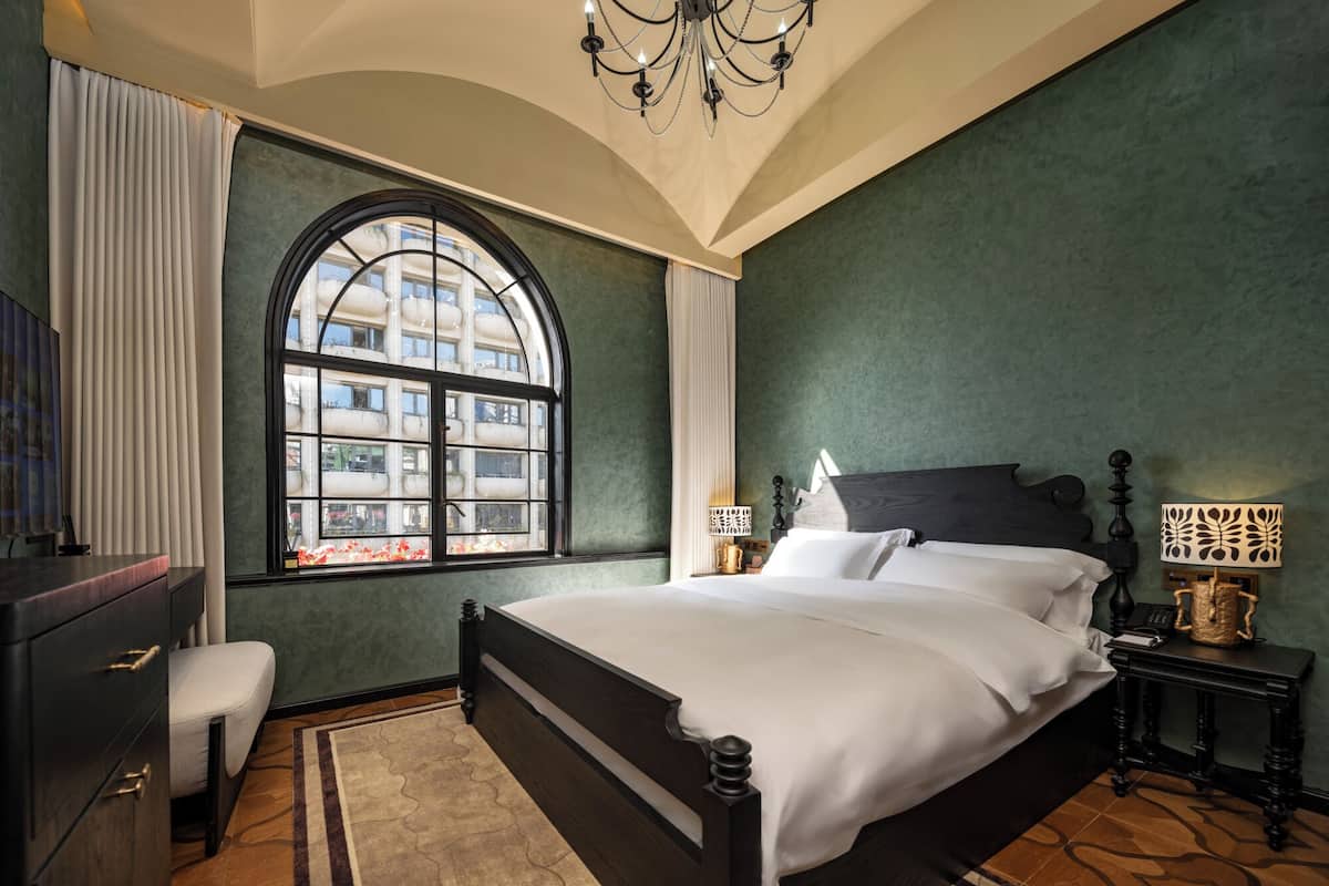A bedroom with a large bed, a dresser, a nightstand, a lamp, and a window with a view of buildings.