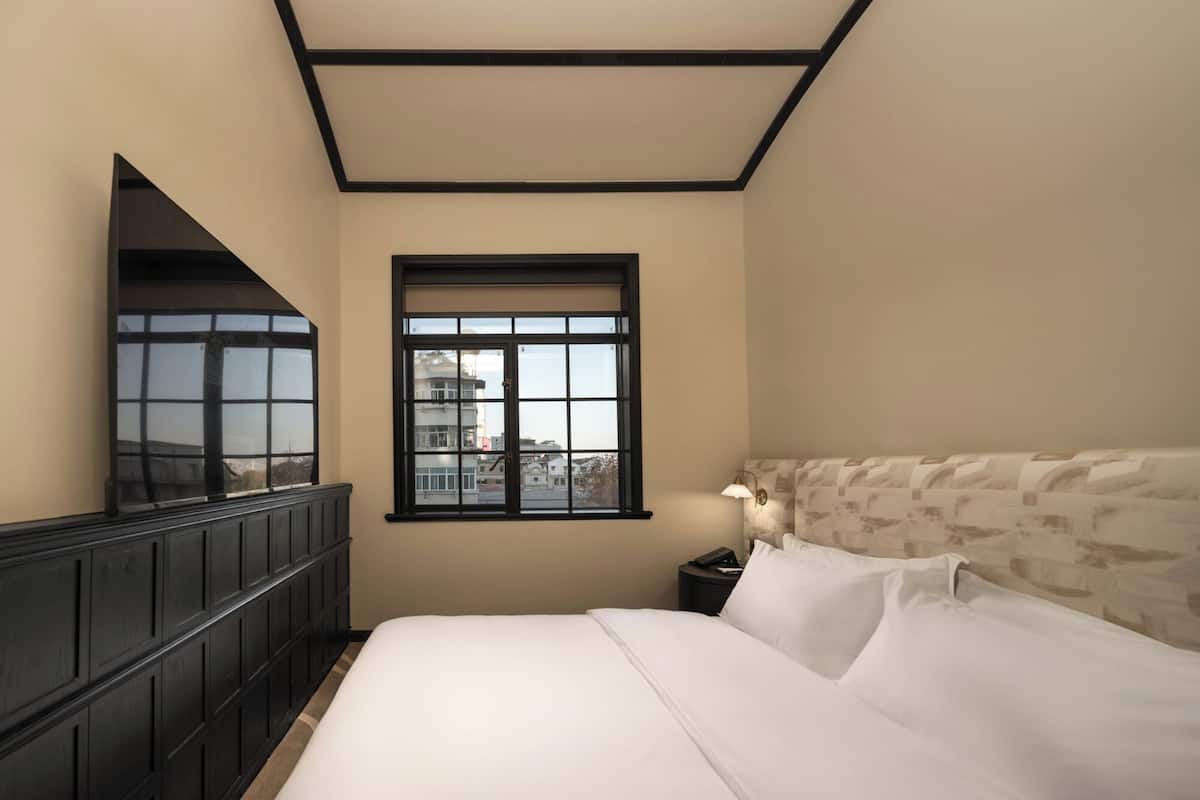 A hotel room with a large bed, a nightstand, a lamp, a window with a city view, and a dark wooden wardrobe.