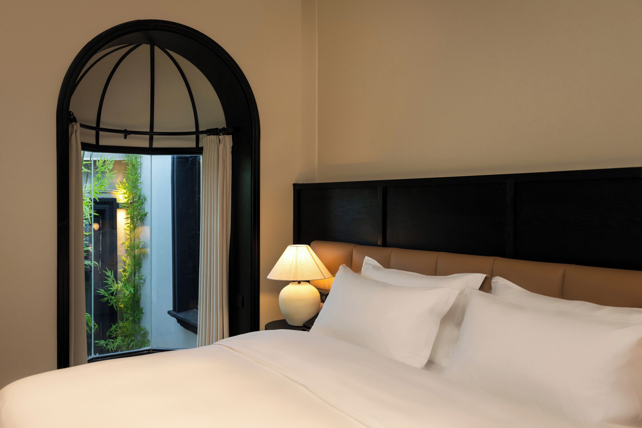 A hotel room with a bed, a nightstand with a lamp, and a view of greenery through an arched window.