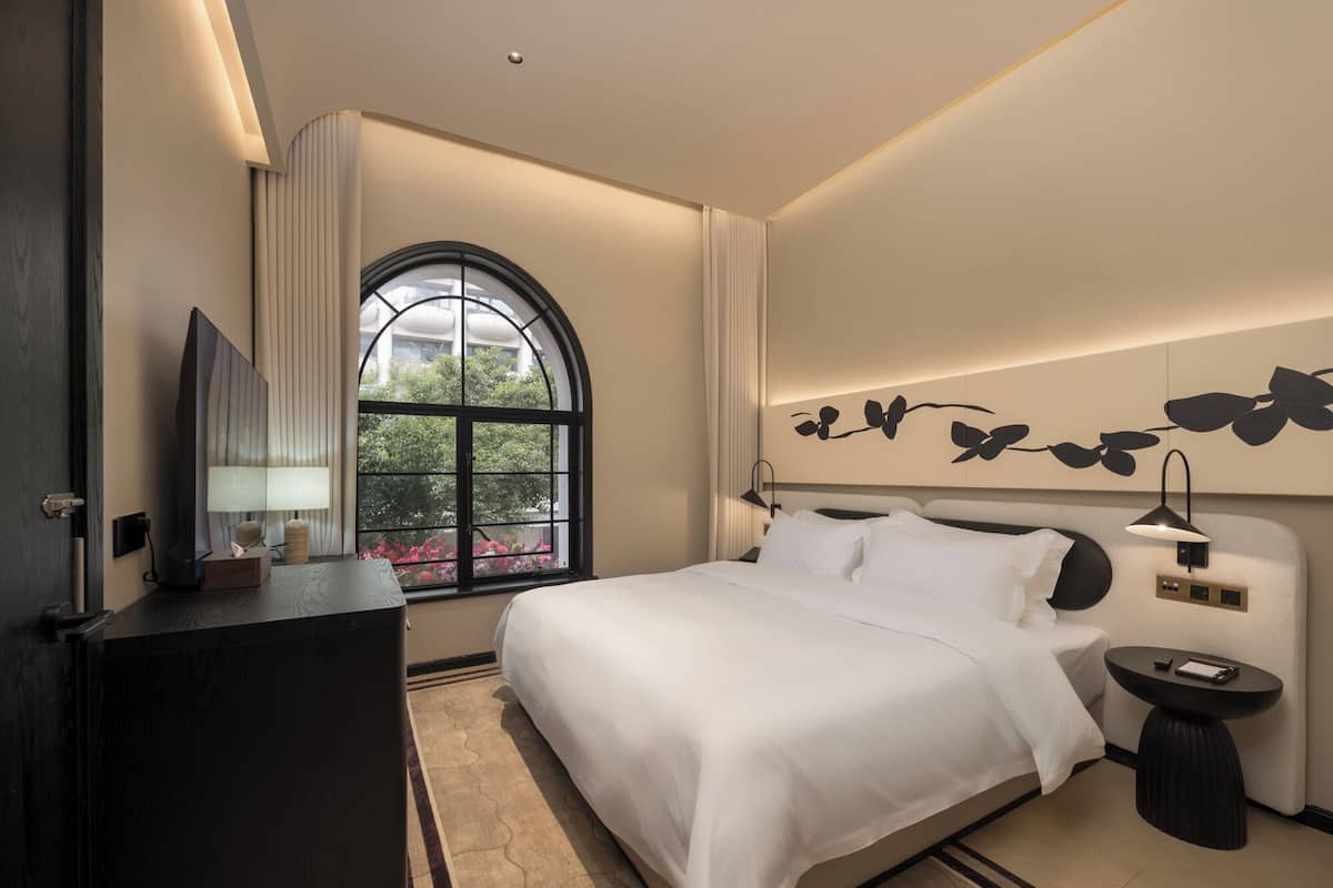 A modern hotel room with a large bed, a bedside table, a nightstand, a mirror, and a window with a view of greenery.