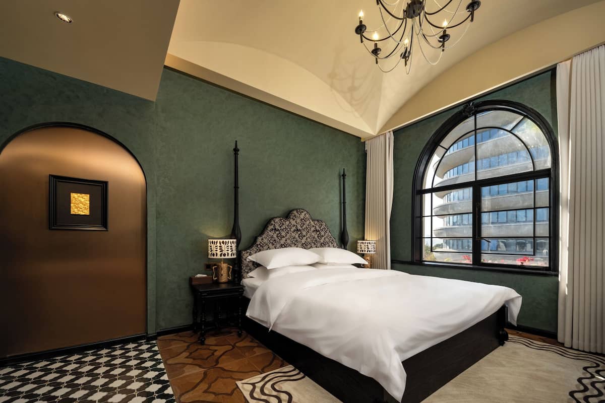 A bedroom with a large bed, a chandelier, a nightstand with a lamp, a framed picture on the wall, and a large window with a view of buildings.