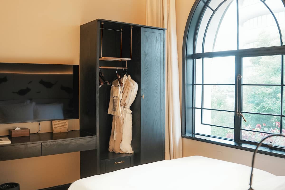 A modern hotel room with a large bed, a wardrobe, a TV, and a window with a view of greenery.