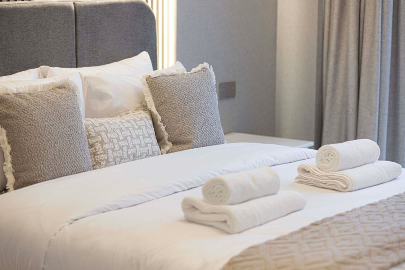 Een net opgemaakt bed met witte lakens en opgerolde handdoeken, een grijs bekleden hoofdbord en een geborduurde plaid.