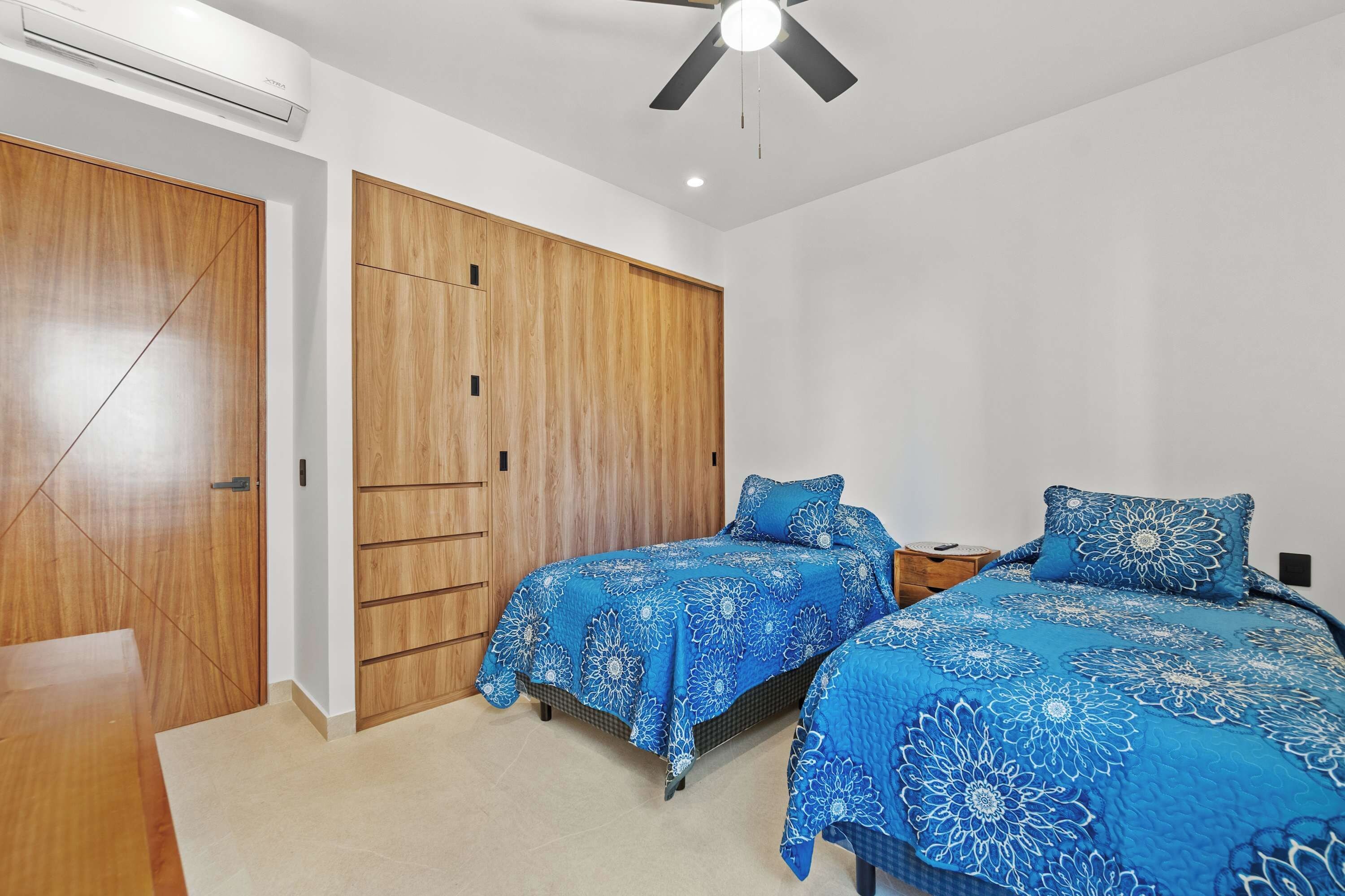 A bedroom with two beds, wooden wardrobe, ceiling fan, and air conditioning unit.
