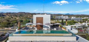 A rooftop pool with lounge chairs and umbrellas, surrounded by a modern building with a view of a hilly landscape.