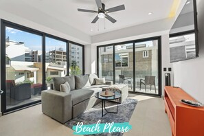 A modern living room with a grey sofa, a small round table, and a ceiling fan. Large windows offer views of the surrounding buildings.