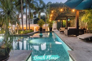 Une piscine dont le bord est revêtu de carreaux, entourée de plantes en pot et de mobilier d’extérieur, située sous une zone abritée illuminée par des guirlandes lumineuses.