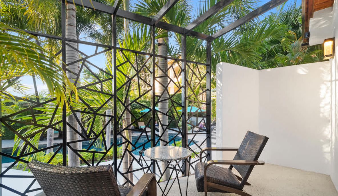 A covered outdoor seating area with a pool in the background, surrounded by palm trees.