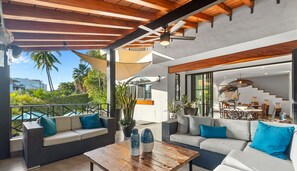 Une vaste terrasse dotée de meubles en osier, d’une table basse en bois et offrant une vue sur la verdure et les bâtiments environnants.