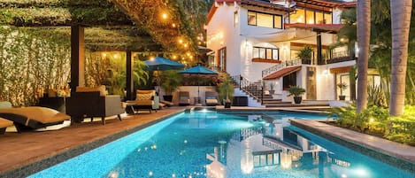 Une piscine entourée de chaises longues et de parasols, elle-même bordée d’une maison dotée d’un balcon et d’un escalier.