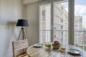 A dining area with a round table, a chair, a lamp, and a view of a building through a large window. - Bright Studio in the Heart of the 13th - Mid-T (Paris)