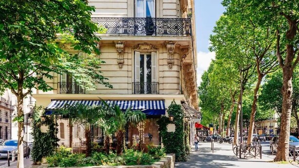 Vista d'un carrer que mostra un edifici amb un balcó, una zona d'estar exterior coberta i una filera d'arbres.